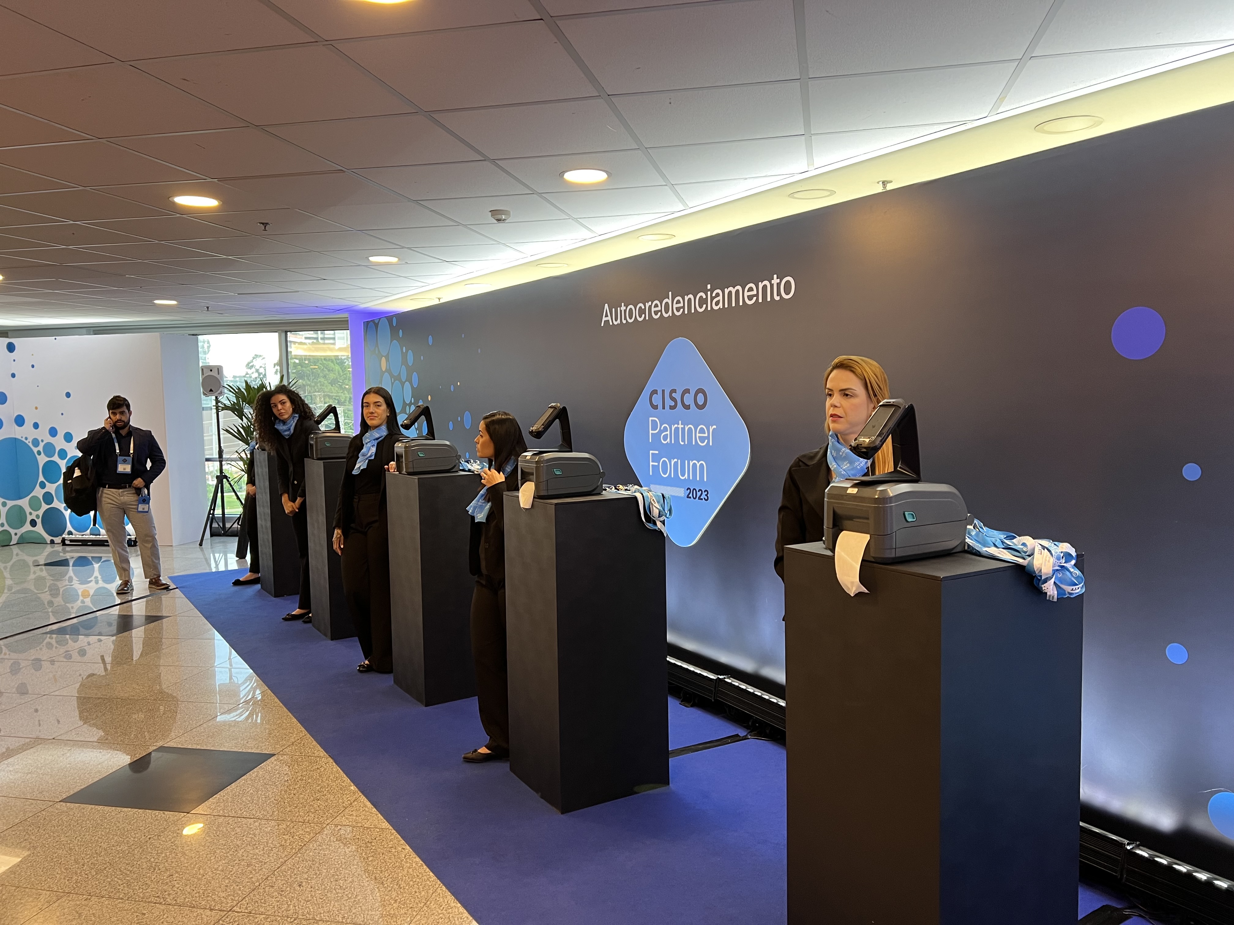 Ilha de autocredenciamento do Grupo INAC no Cisco Partner Forum 2023, apresentando totens de autoatendimento minimalistas com scanners de alta performance e impressoras térmicas integradas.