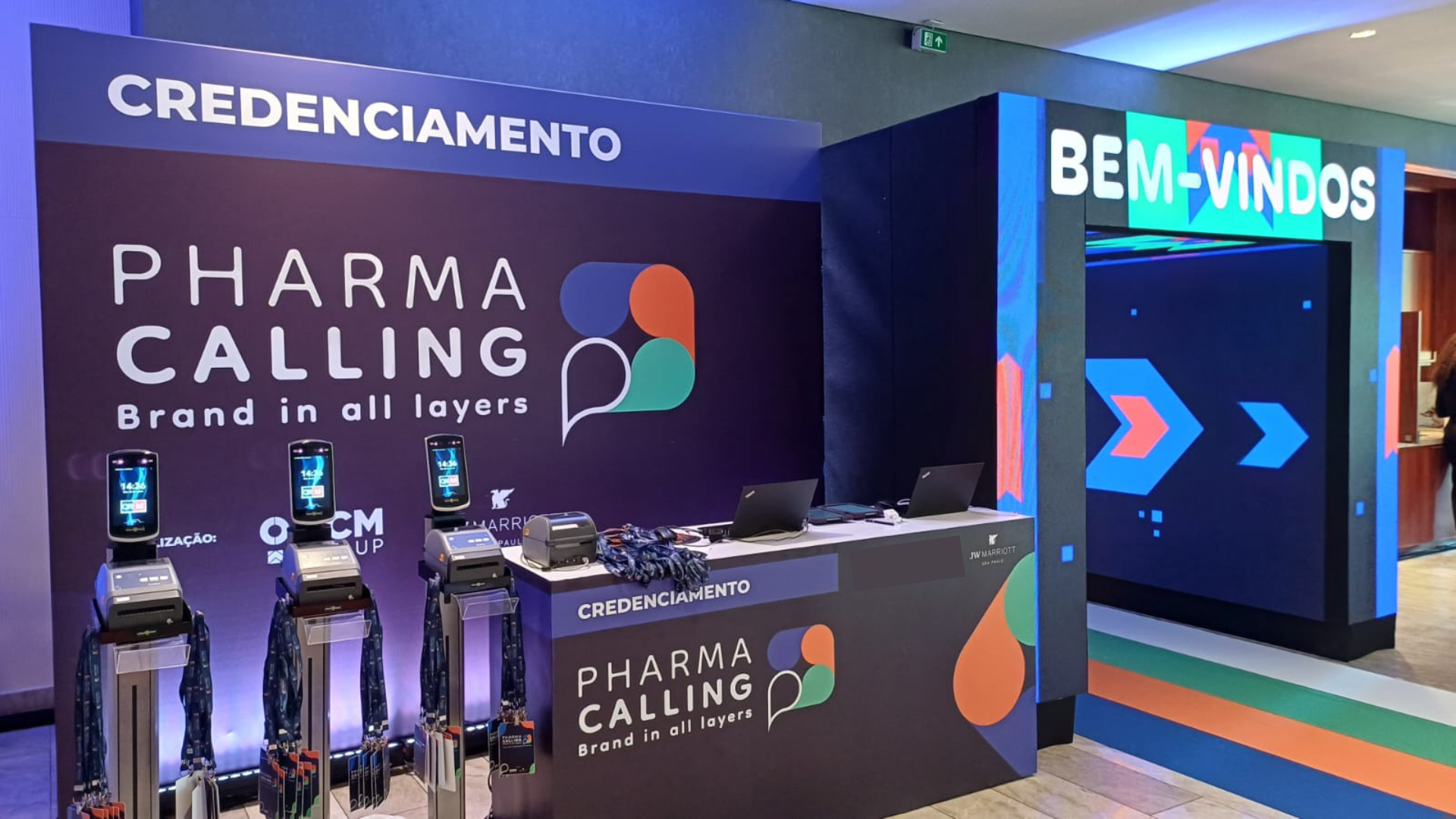 Estrutura de credenciamento tecnológico no evento Pharma Calling, com totens de reconhecimento facial e impressoras térmicas integradas em um balcão moderno com portal de LED.