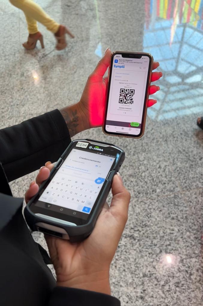 Close de atendente utilizando coletor de dados Zebra com scanner laser vermelho para ler QR Code de ingresso no smartphone de um participante em evento.