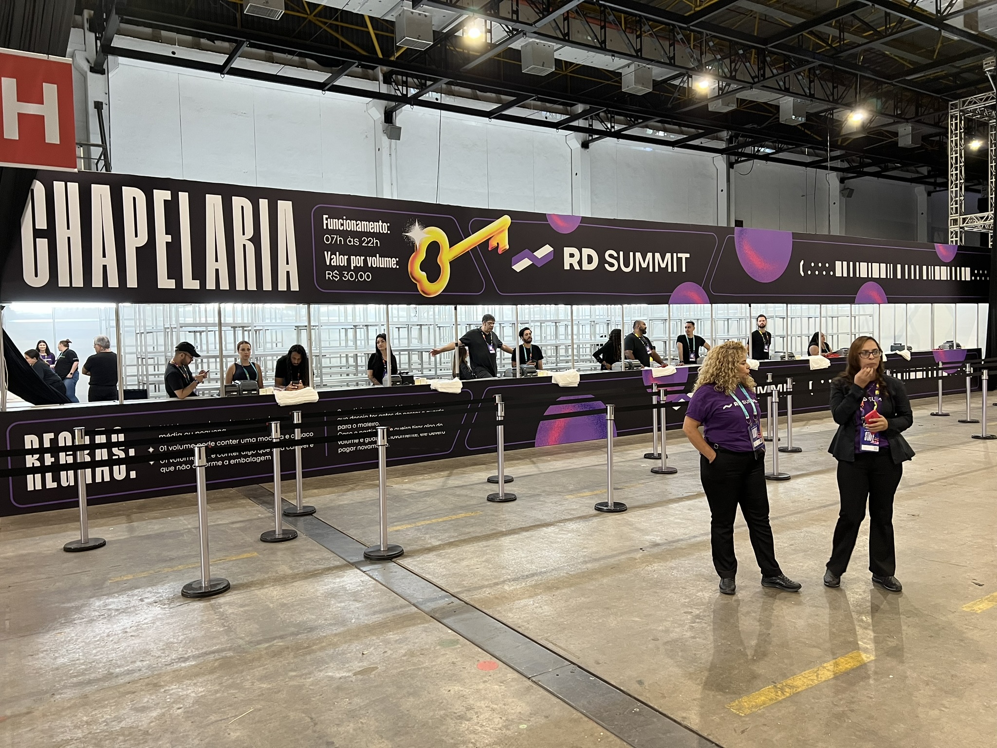 Operação de chapelaria e guarda-volumes digital do Grupo INAC no evento RD Summit, exibindo balcão de atendimento organizado com estantes ao fundo e sinalização profissional.